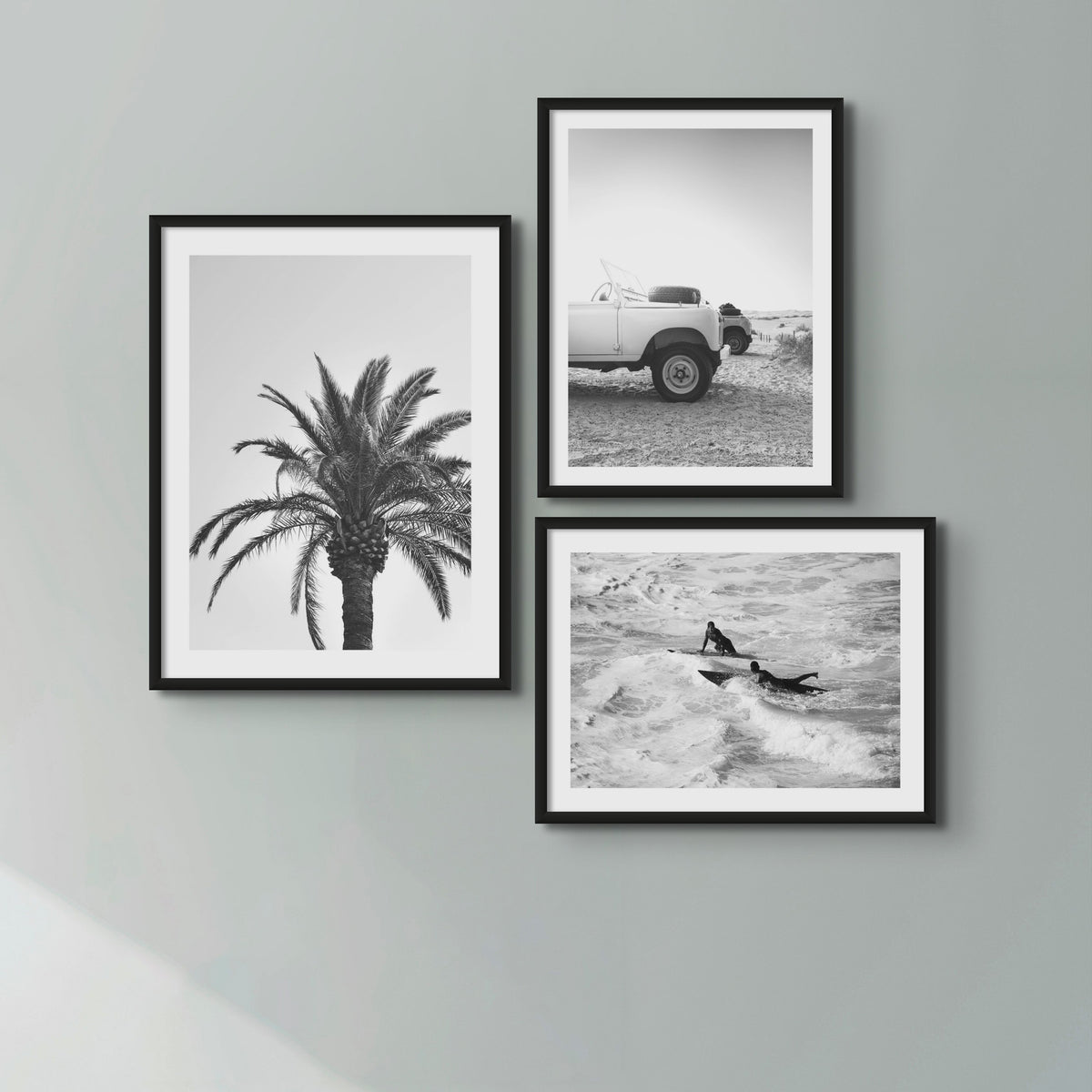 Three black and white photographs of a palm tree, a Land Rover Defender and two men on surf boards paddling out through the waves. The photographs are in black frames on a grey wall. 