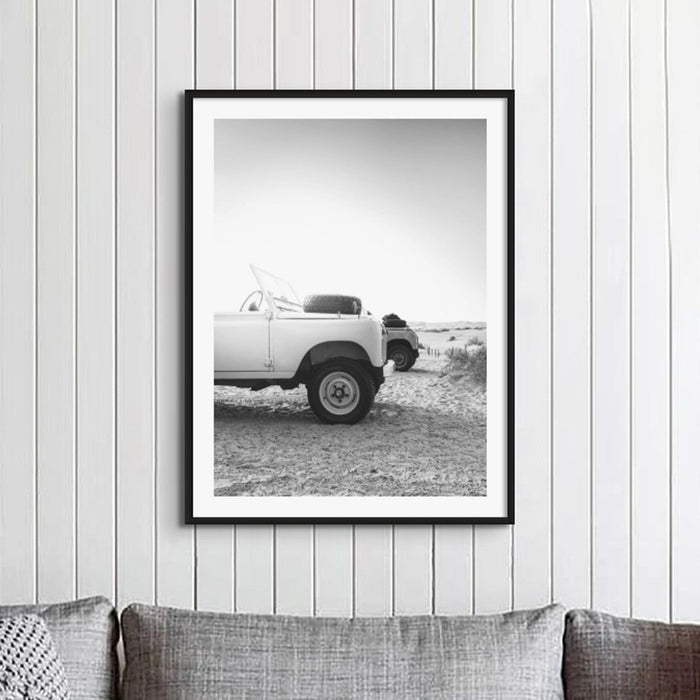 Black and white photograph of Land Rover Defender on a beach at sunrise on a timber panelled wall in a loungeroom above a grey fabric sofa with cushions