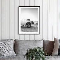 Black and white photograph of Land Rover Defender on a beach at sunrise on a timber panelled wall in a loungeroom above a grey fabric sofa with cushions