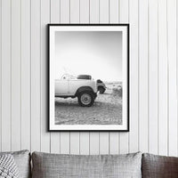 A black and white photograph of two land rover defenders on a beach in the early morning. The monochrome photograph is in a black frame