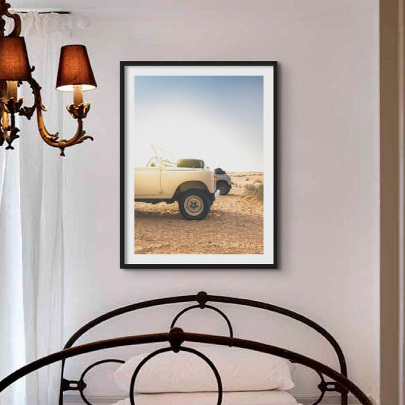 a photograph of land rover defenders on the beach ealry in the morning, in a black frame above a victorian bed in a small bedroom with a victorian chandelier