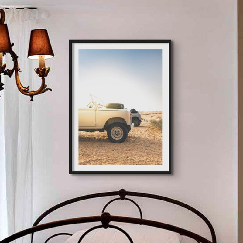 A faded colour photograph of land rover defenders on a beach in the early morning in a black frame. The photograph is above a small victorian bed with wrought iron bedhead, in a small room with a victorian chandelier.