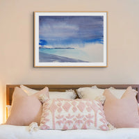 blue seascape painting framed, with headland and beach in foreground. the watercolour beach paitning is framed in a natural wood frame and is above a bed